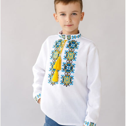 A boy wearing a white embroidered shirt with the Ukrainian coat of arms, featuring blue and yellow embroidery, a yellow tassel on the collar, and the shirt is complemented by decorative tassels on the collar and cuffs.